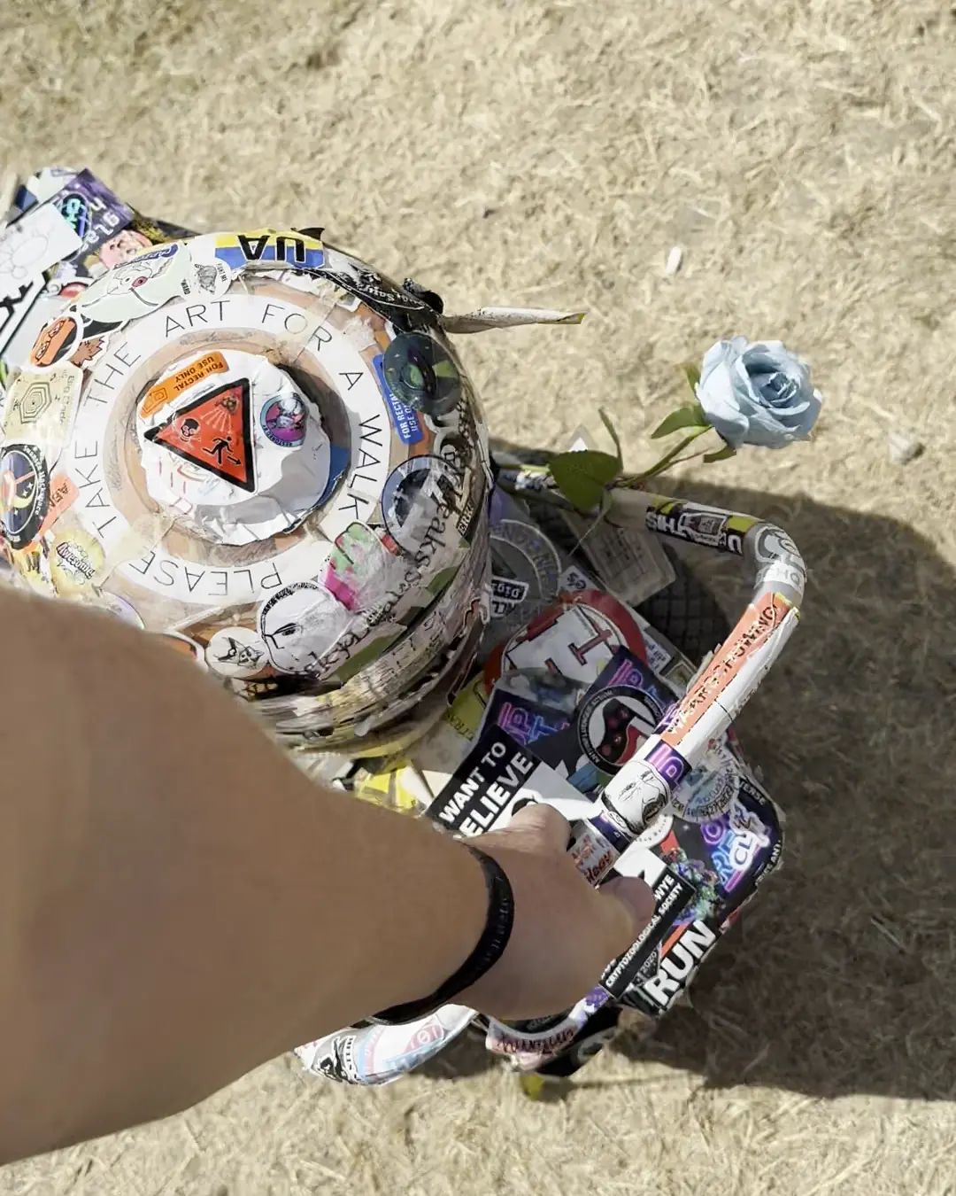Looking down at a sticker-covered trolley with a blue rose tucked into it and "Please take the art for a walk" written round the top, my hand on the handle
