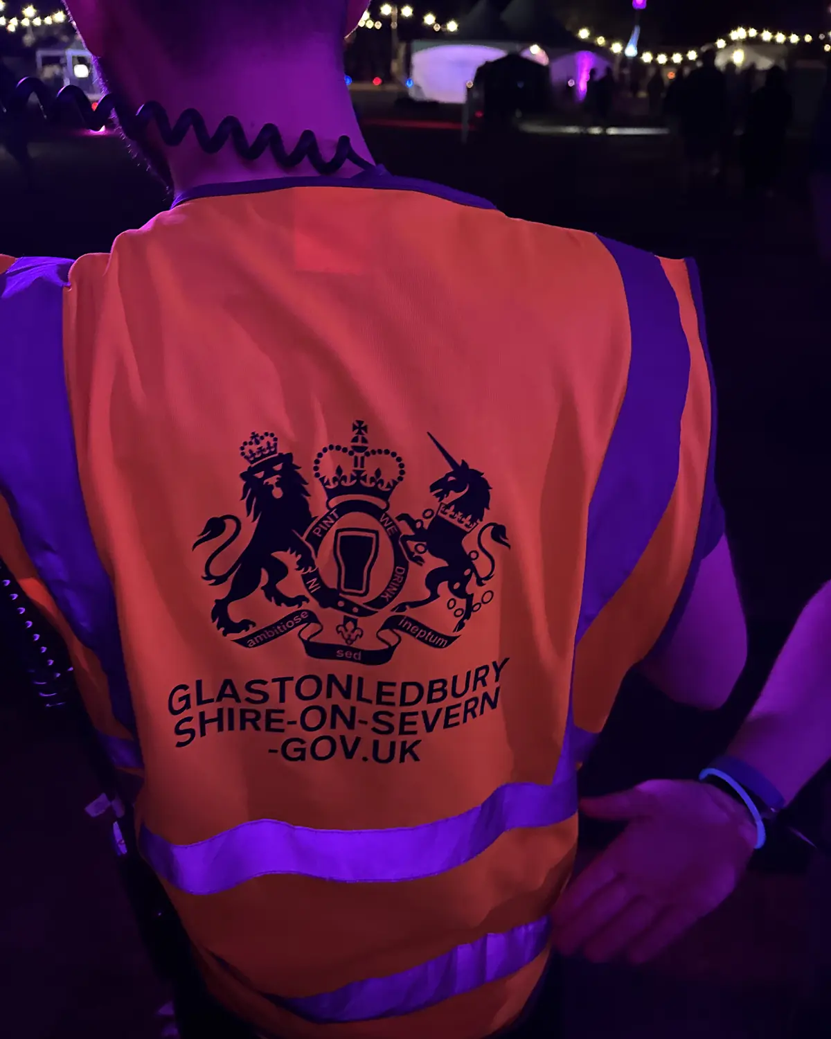 The back of a hi-vis vest at night, printed with a mock coat of arms that says "In pint we drink" and "Glastonledbury Shire-on-Severn -gov.uk"