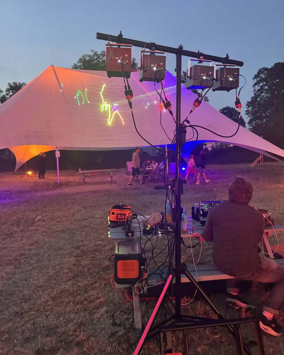 4 laser projectors on a stand drawing glowing figures onto a big white tent at dusk, run from a laptop on a picnic table