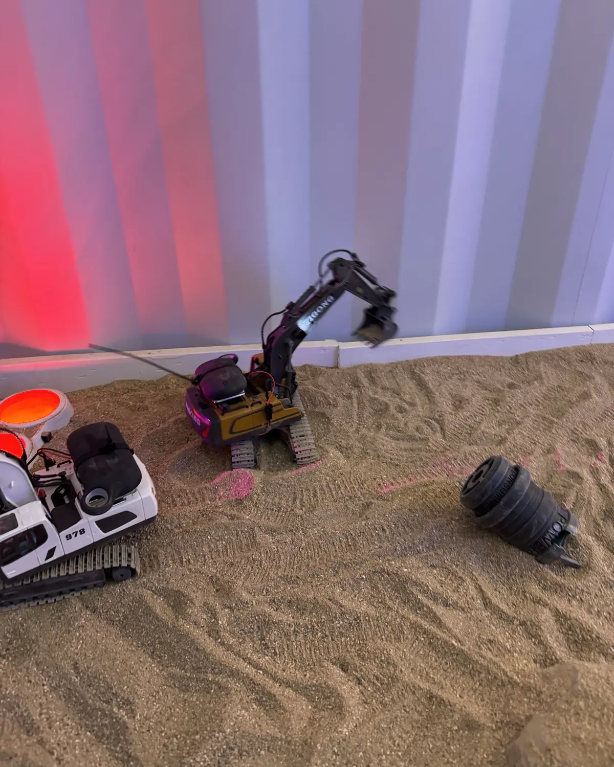 Remote-controlled diggers in a sandpit, lit red and blue against a white wall
