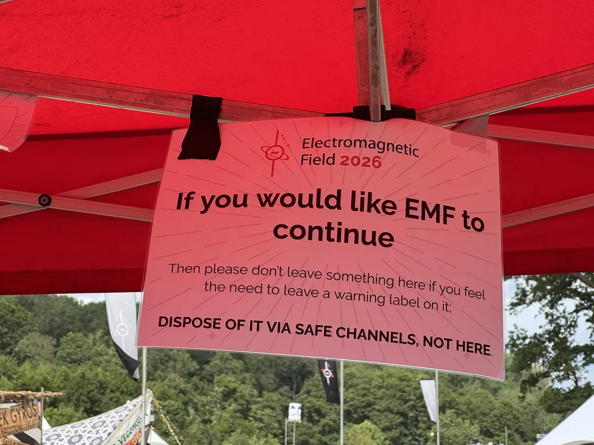 An EMF 2026 sign under a red gazebo: "If you would like EMF to continue, then please don't leave something here if you feel the need to leave a warning label on it; dispose of it via safe channels, not here."